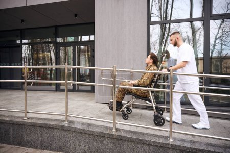 Photo for Side view of hospital attendant pushing wheelchair with serviceman towards medical facility front door - Royalty Free Image