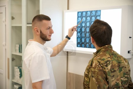 Profesional médico señalando con lápiz en la imagen de la resonancia magnética cerebral al militar en su oficina