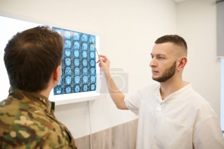 Seriöser Neurologe zeigt mit Bleistift auf Gehirn-MRT-Bild zu Soldat in seinem Büro