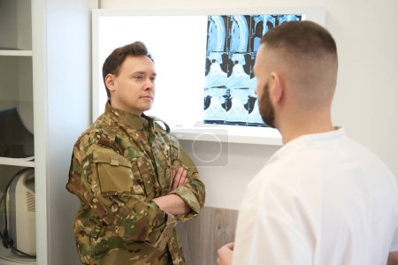 Serviceperson with arms crossed standing in front of negatoscope with thoracic spine CT images during conversation with traumatologist