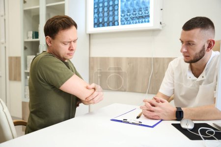 Téléchargez les photos : Triste homme adulte touchant son coude assis au bureau devant le médecin - en image libre de droit