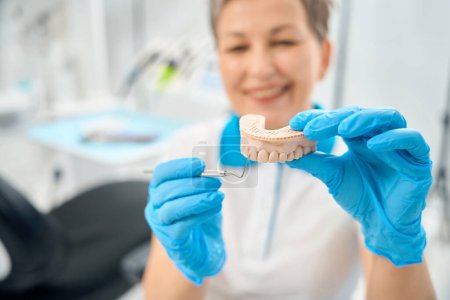 Photo for Smiling woman dental and aesthetic practitioner holding 3d model of upper jaw and pointing with sickle probe to foretooth, explaining way of reconstruction, tooth veneer - Royalty Free Image