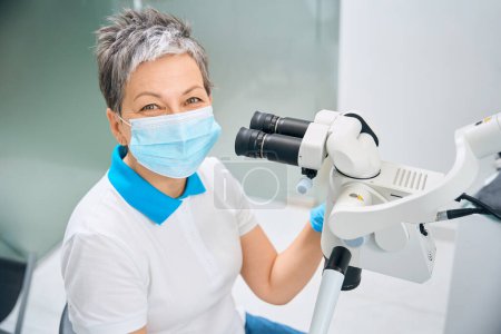 Photo for Female laboratory technician or doctor assistant in protective face mask sitting near big microscope, carrying out precisely treatment or preparing implants or braces before installation - Royalty Free Image