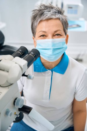 Foto de Mujer con estilo y positivo odontólogo sentado en su oficina cerca del microscopio y sonriendo bajo la máscara protectora, haciendo su trabajo con precisión y precisión - Imagen libre de derechos
