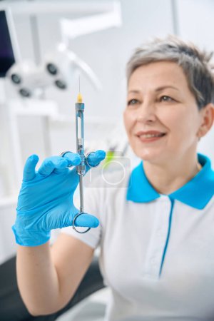 Foto de Enfermera sonriente de terapeuta dental sosteniendo jeringa de vidrio con anestésico en el interior, alivio del dolor antes de comenzar el chequeo y tratamiento de los dientes, enfoque de cuidado a los pacientes - Imagen libre de derechos