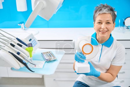 Foto de Mujer profesional dental calificada sonriente sentada en su lugar de trabajo cerca de herramientas dentales y sosteniendo en las manos un dispositivo de rayos X dental portátil - Imagen libre de derechos
