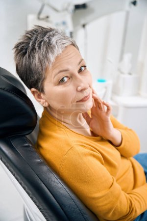 Téléchargez les photos : Femme âgée touchant la joue douloureuse assise sur une chaise d'examen dans un cabinet de thérapeute dentaire, ressentant une sensibilité dentaire en raison d'une maladie des gencives ou des gencives, d'un émail cassé - en image libre de droit