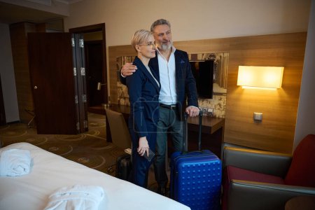 Adult caucasian businessman with suitcase hugging businesswoman and they looking away in hotel room at daytime. Concept of business trip, vacation and travelling. Idea of teamwork