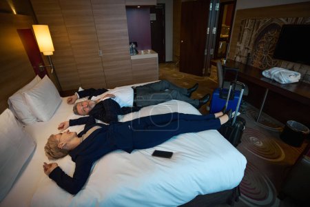 High angle view of serious adult caucasian businessman and smiling businesswoman lying on bed and looking at each other in hotel room at daytime. Concept of business trip, vacation and travelling