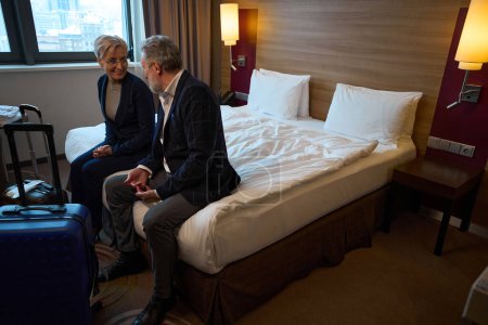 Adult caucasian businessman and smiling businesswoman sitting on bed and looking at each other in hotel room at daytime. Concept of business trip, vacation and travelling. Idea of teamwork