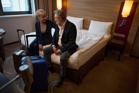 Middle aged caucasian businessman and smiling businesswoman sitting on bed and looking at each other in hotel room at daytime. Concept of business trip, vacation and travelling. Idea of teamwork