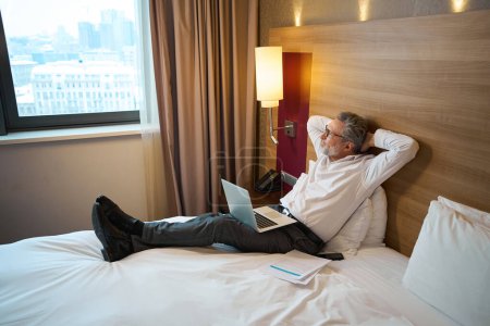 Foto de Pensativo adulto hombre de negocios caucásico con portátil mirando hacia otro lado, acostado en la cama en la habitación de hotel durante el día. Concepto de viaje de negocios, vacaciones y viajes - Imagen libre de derechos