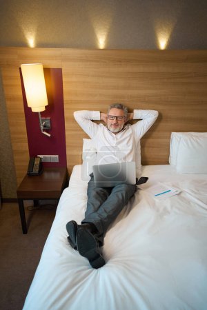 Photo for Smiling adult caucasian businessman with laptop looking at camera, lying on bed in hotel room at daytime. Concept of business trip, vacation and travelling - Royalty Free Image