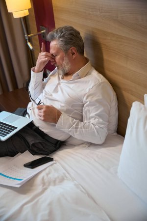 Téléchargez les photos : Vue grand angle fatigué homme d'affaires caucasien adulte pendant le travail sur le lit dans la chambre d'hôtel le jour. Concept de voyage d'affaires, vacances et voyages - en image libre de droit