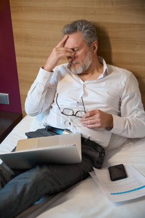 Téléchargez les photos : Vue en angle élevé épuisé adulte caucasien entrepreneur masculin pendant le travail sur le lit dans la chambre d'hôtel le jour. Concept de voyage d'affaires, vacances et voyages - en image libre de droit