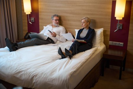 Téléchargez les photos : Homme d'affaires caucasien adulte regardant une femme d'affaires concentrée regardant des documents sur le lit dans une chambre d'hôtel le jour. Concept de voyage d'affaires, vacances et voyages. Idée de travail d'équipe - en image libre de droit