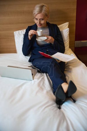 Téléchargez les photos : Vue en angle élevé d'une femme d'affaires caucasienne adulte concentrée buvant du thé ou du café dans une tasse et regardant un ordinateur portable sur son lit dans une chambre d'hôtel le jour. Concept de voyage d'affaires, vacances et voyages - en image libre de droit