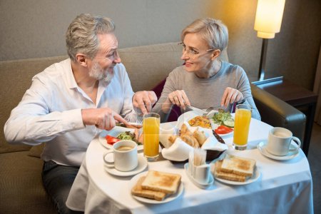 Téléchargez les photos : Couple caucasien adulte souriant prenant le petit déjeuner et se regardant dans la chambre d'hôtel le matin. Concept de repos, vacances et voyages. Idée de relation - en image libre de droit