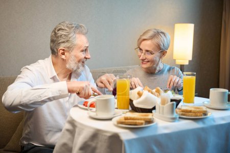 Téléchargez les photos : Couple caucasien mature souriant petit déjeuner et se regardant dans la chambre d'hôtel le matin. Concept de repos, vacances et voyages. Idée de relation - en image libre de droit