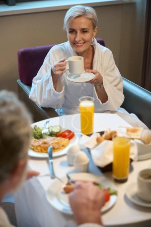 Téléchargez les photos : Femme caucasienne adulte souriante buvant du thé ou du café dans une tasse et regardant un homme partiel prendre le petit déjeuner dans une chambre d'hôtel le matin. Concept de repos, vacances et voyages. Idée de relation - en image libre de droit