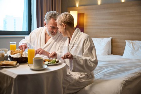 Téléchargez les photos : Heureux couple caucasien mature prenant le petit déjeuner et se regardant sur le lit à table dans la chambre d'hôtel le matin. Concept de repos, vacances et voyages. Idée de relation - en image libre de droit
