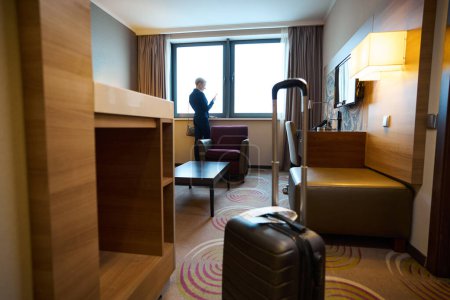 Photo for Distance view of businesswoman watching smartphone by window in hotel room with suitcase at daytime. Concept of business trip, vacation and travelling - Royalty Free Image