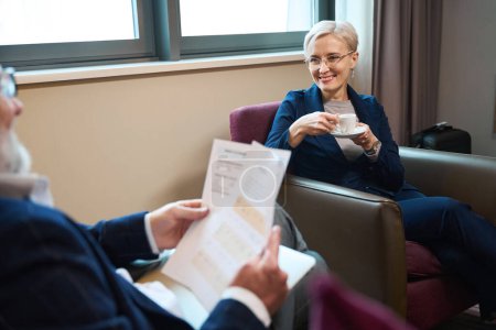 Photo for Smiling caucasian businesswoman drinking tea or coffee and looking at blurred cropped businessman with laptop and documents in hotel room at daytime. Business trip, vacation and travelling. Teamwork - Royalty Free Image