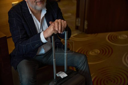 Foto de Cara oscura del hombre de negocios barbudo con maleta sentada en una silla de cuero en una habitación de hotel borrosa durante el día. Concepto de viaje de negocios, vacaciones y viajes - Imagen libre de derechos