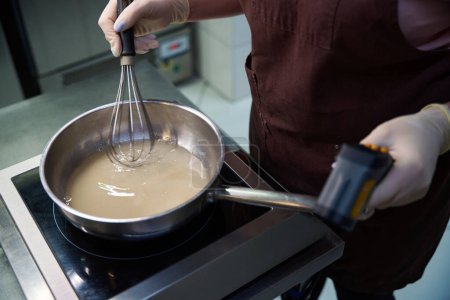 Qualifizierte Konditorin rührt Geliermittel Agar-Agar-Mischung in Topf bei mittlerer Hitze bis zum Kochen, Temperatur mit professionellem Küchenthermometer überprüfen