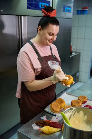 Mujer sonriente pastelera sosteniendo bolsa de pastelería con natillas y llenando las crocantes y sabrosas chouquetas con relleno dulce, dulces