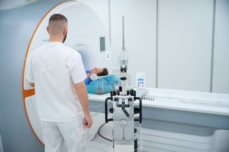 Téléchargez les photos : Technologue MR opérant une table d'IRM motorisée avec patient dans un casque antibruit couché en position couchée - en image libre de droit