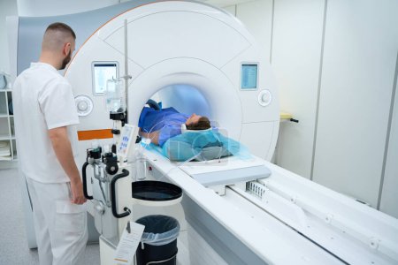 Röntgengerät drückt Taste auf MRT-Portalsteuerung, während Patient mit Kopfhörer und Spule um Bein auf Scan-Tisch liegt