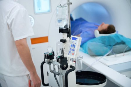 Ausgeschnittenes Foto eines Röntgenbildners, der vor dem Kontrastinjektor steht, während der Patient mit geräuschunterdrückenden Kopfhörern auf dem MRT-Tisch liegt