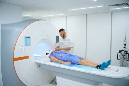 Foto de Hombre adulto acostado supino en la mesa de imágenes de resonancia magnética mientras radiografo coloca la bobina sobre su cabeza - Imagen libre de derechos