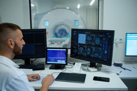 Röntgengerät sitzt am Schreibtisch im Kontrollraum und betrachtet die Magnetresonanztomographie des Gehirns des Patienten, die auf dem Computermonitor angezeigt wird