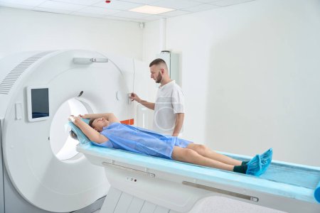 Foto de Paciente con los brazos levantados por encima de la cabeza acostado en la mesa CT mientras el radiografo presiona el botón en el panel de control del pórtico - Imagen libre de derechos