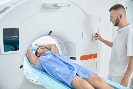 Mann mit über Kopf erhobenen Armen auf Computertomographietisch liegend, während Radiologe Knopf an Portalsteuerung drückt
