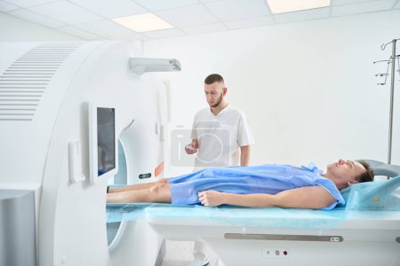 Man lying supine while radiologic technologist moving examination table into CT gantry aperture with hand-held controller
