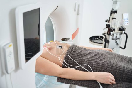 Ausgeschnittenes Foto des Patienten mit EKG-Elektroden auf der Brust liegend auf dem Untersuchungstisch in der CT-Portalöffnung
