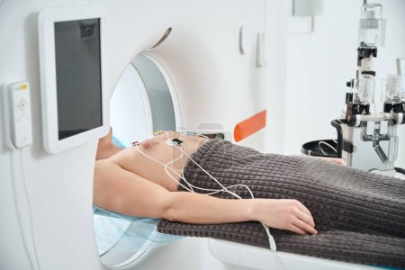Ausgeschnittenes Foto eines Mannes mit EKG-Elektroden, die an der Brust befestigt sind, liegend auf dem Untersuchungstisch in der Computertomographie-Portalöffnung