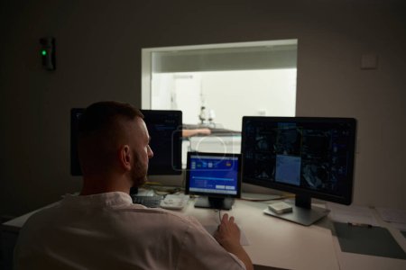 Profesional de la salud sentado en el escritorio en la sala de control mientras mira las exploraciones por TC en el monitor de la computadora