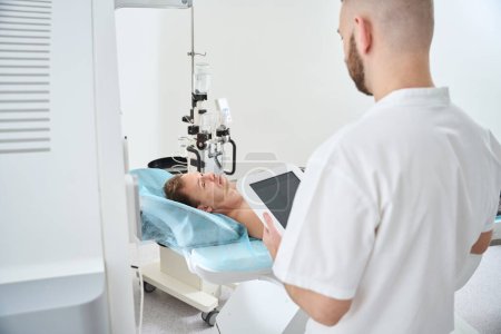 Tecnólogo radiológico con tableta en las manos de pie frente al hombre acostado en la mesa de tomografía computarizada cerca del inyector de contraste