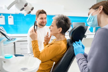 Foto de Terapeuta dental sonriente y su asistente femenina mirando a la cliente mujer satisfecha disfrutando de su sonrisa blanca saludable en el reflejo del espejo, atención dental de alta calidad - Imagen libre de derechos