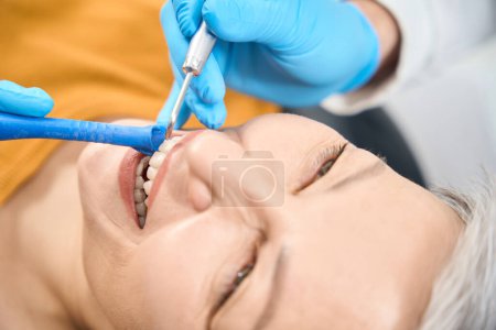Photo for Close-up mature lady smiling visiting aesthetic dentist, doctor and assistant using combination of water, compressed air and fine powder particles of sodium bicarbonate to clean and polish the teeth - Royalty Free Image