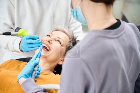 Foto de Paciente anciana sonriendo mientras el dentista calificado y su asistente mantienen fuera el procedimiento de limpieza y pulido de dientes de flujo de aire, mejora de brillo dental - Imagen libre de derechos