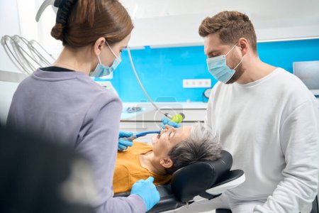 Téléchargez les photos : Vue du dentiste assistant tenant l'éjecteur de salive pendant le thérapeute dentaire qualifié gardant le traitement de flux d'air des dents matures du patient, le nettoyage et le polissage, l'élimination de la souche - en image libre de droit
