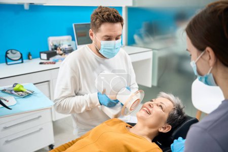 Téléchargez les photos : Dentiste esthétique dans un masque protecteur tenant une radiographie dentaire portable près d'une patiente, faisant l'image de dents pour vérifier les zones problématiques et trouver des obturations cassées ou des dents avec carie - en image libre de droit
