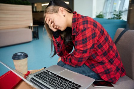 Téléchargez les photos : Jeune asiatique épuisée employée des TI avec migraine toucher sa tête pendant le travail au bureau de coworking. Concept de travail indépendant ou à distance moderne - en image libre de droit