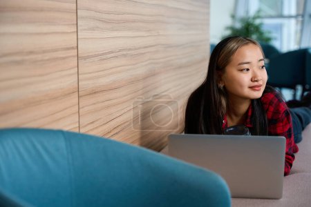 Téléchargez les photos : Jeune asiatique souriante employée de l'informatique avec café regardant loin tout en travaillant sur ordinateur portable sur canapé dans le bureau de coworking. Concept de travail indépendant ou à distance moderne - en image libre de droit