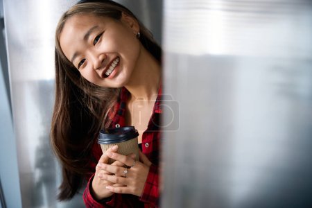 Téléchargez les photos : Partielle jeune asiatique souriante gestionnaire informatique avec tasse à café regardant la caméra dans le bureau de coworking flou. Concept de travail indépendant ou à distance moderne - en image libre de droit